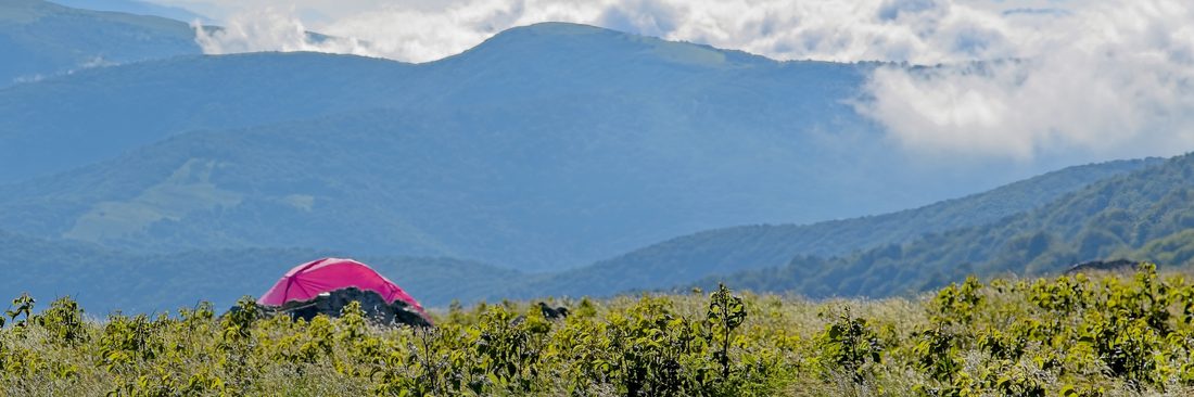 Pink tent perched on a grassy bald with low clouds drifting through distant blue peaks