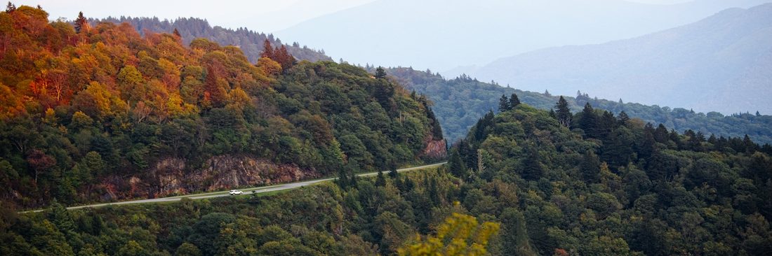 Lone car on a winding Blue Ridge Parkway road beside autumn ridges fading into hazy blue distance