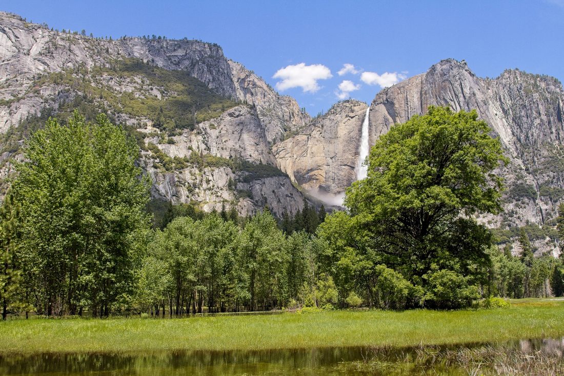 Tall waterfall tumbling down granite cliffs above a sunlit meadow and reflecting pond