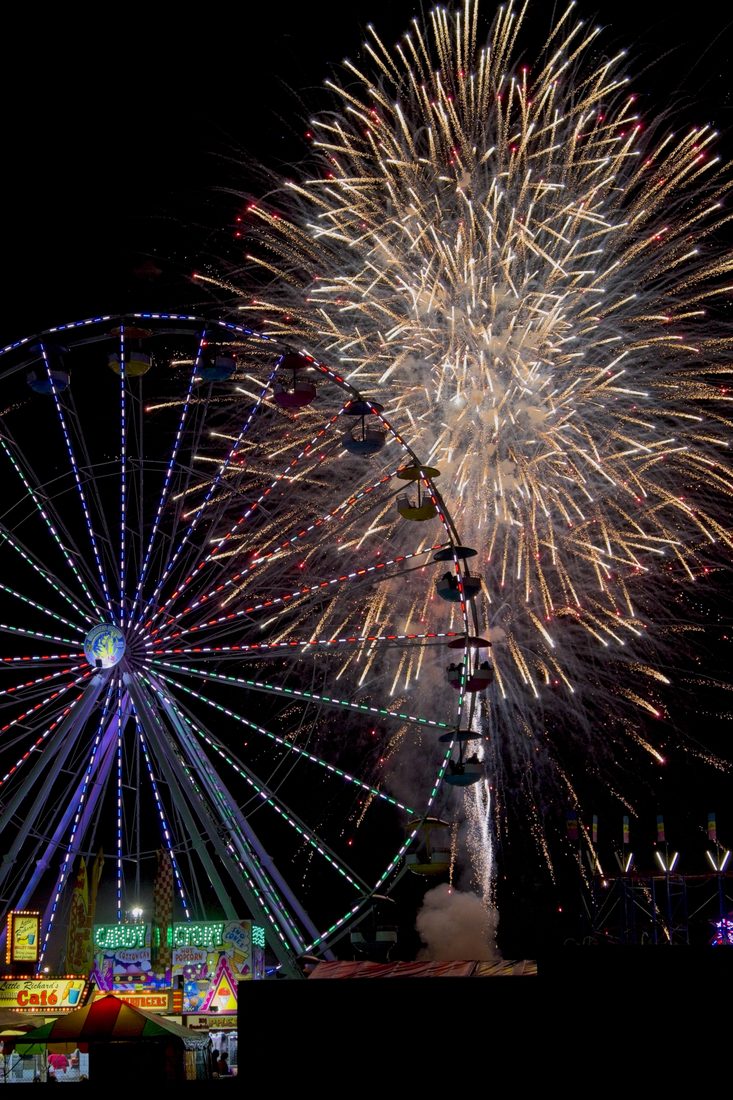 Golden fireworks erupting over a lit ferris wheel and carnival midway at night