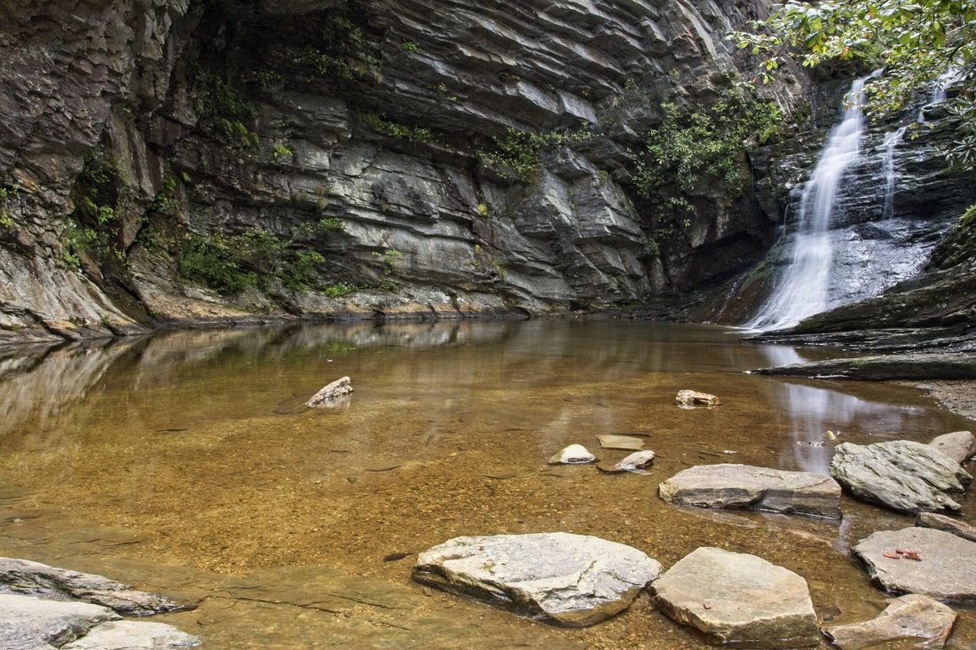 Hidden grotto waterfall spilling into shallow amber pool framed by towering rock cliff